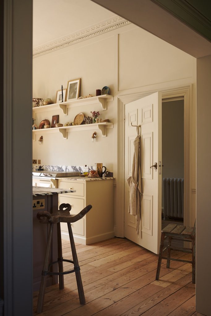 posttitle | sitetitle Light filled vintage kitchen with cream cabinets open shelves of pottery and framed photos and a white door ajar